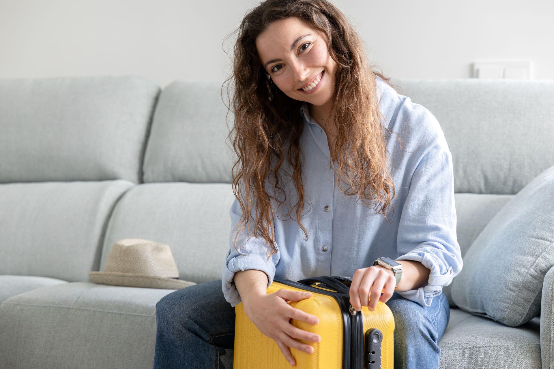  Frau sitzt auf einem Sofa und packt einen gelben Koffer für den Urlaub. Ein Strohhut liegt daneben.