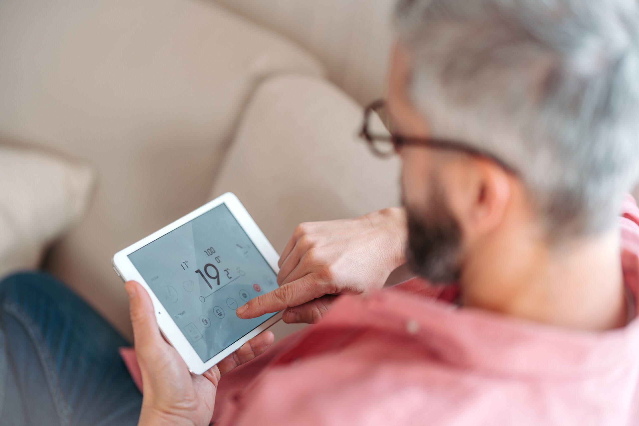 Eine Person sitzt auf einem Sofa mit einem Tablet und bedient eine Heizung 